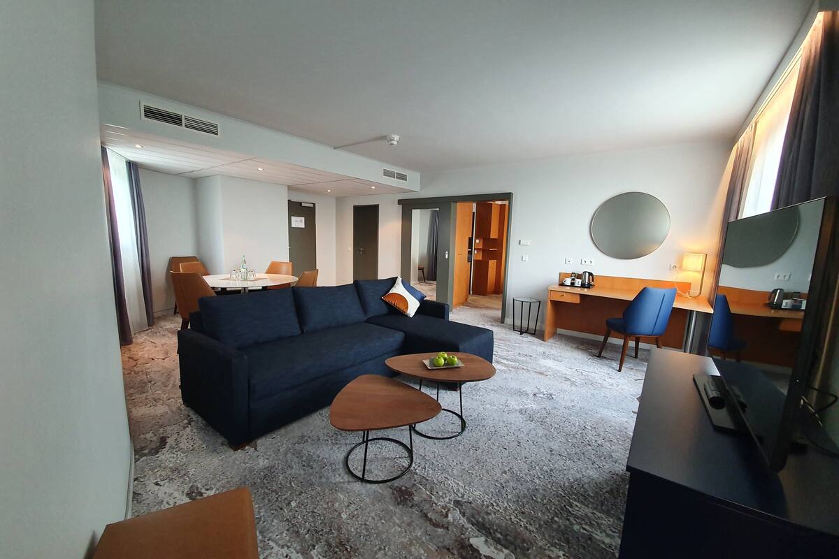 A modern hotel room with a dark blue sofa, a small round table with apples, a desk with a chair, a TV, and a large mirror.