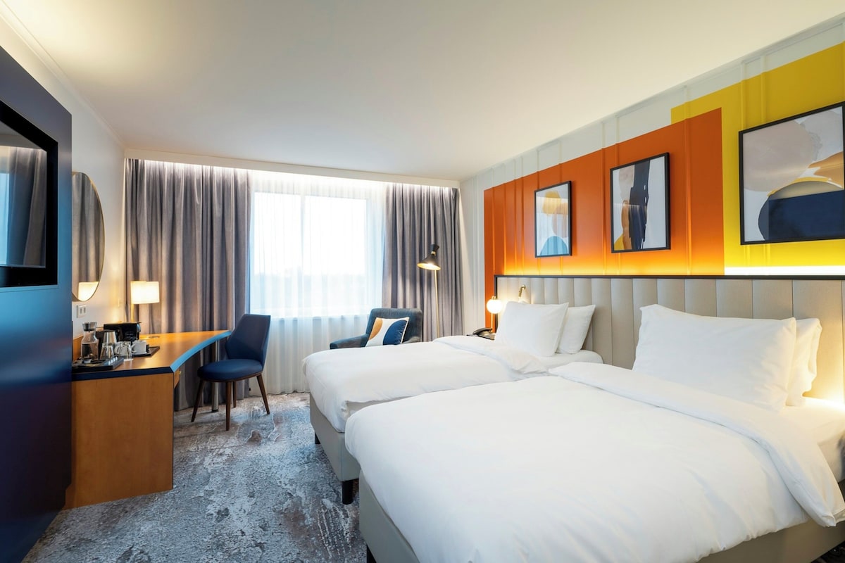 A hotel room with two beds, a desk, a chair, a TV, and a window with curtains.