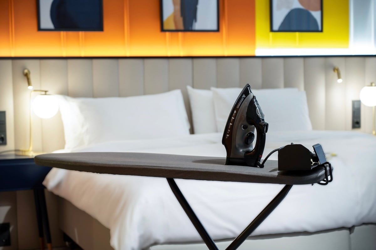 A neatly made bed with white linens, an ironing board with an iron, and a bedside lamp.