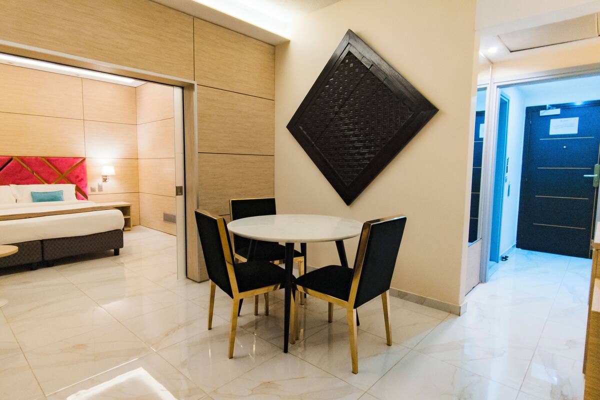 A modern hotel room with a bed, a dining table with chairs, a wall-mounted artwork, and a door leading to another room.