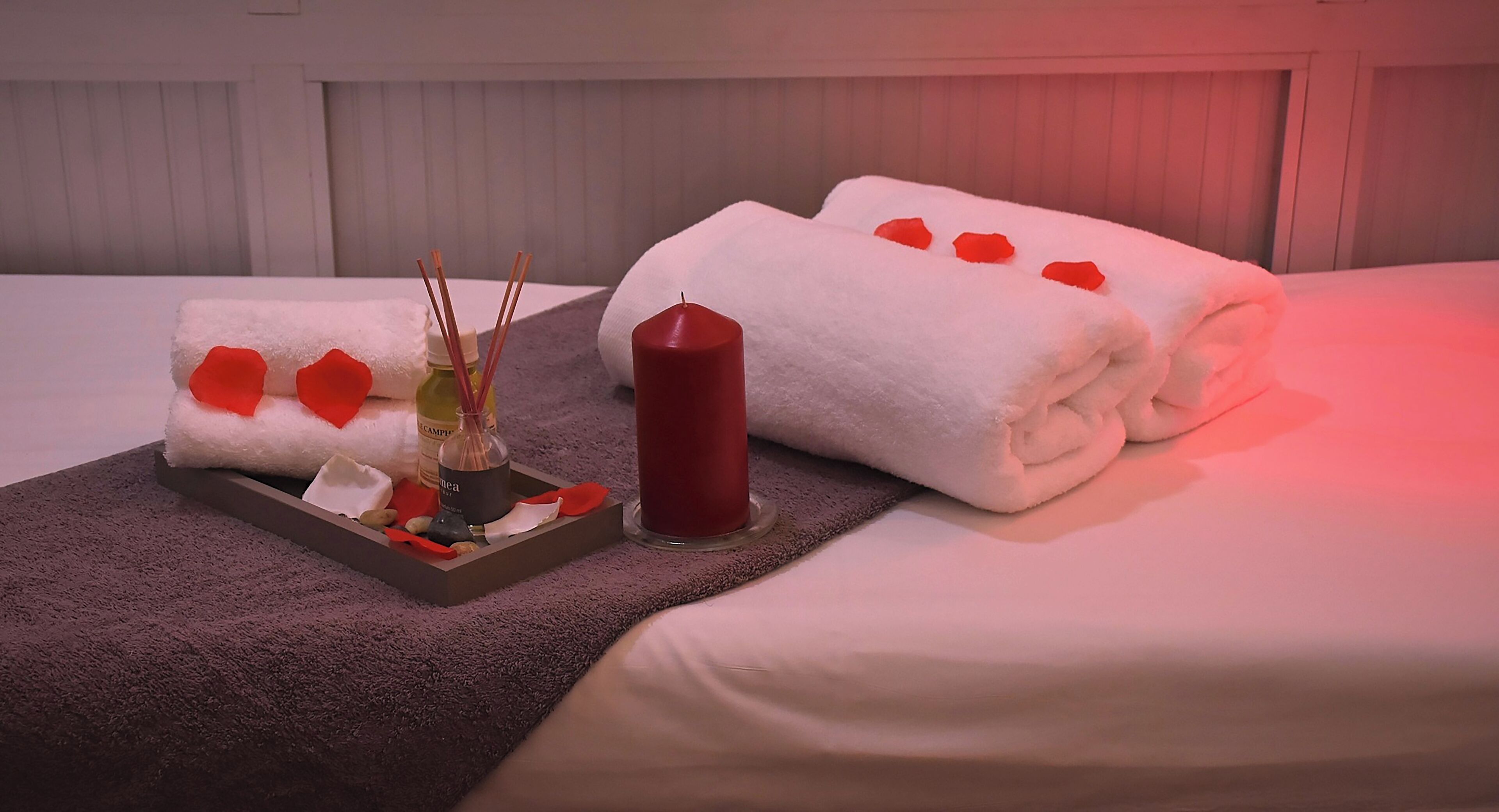 A neatly made bed with white linens and red rose petals, a candle, and a tray with a bottle and reed sticks.