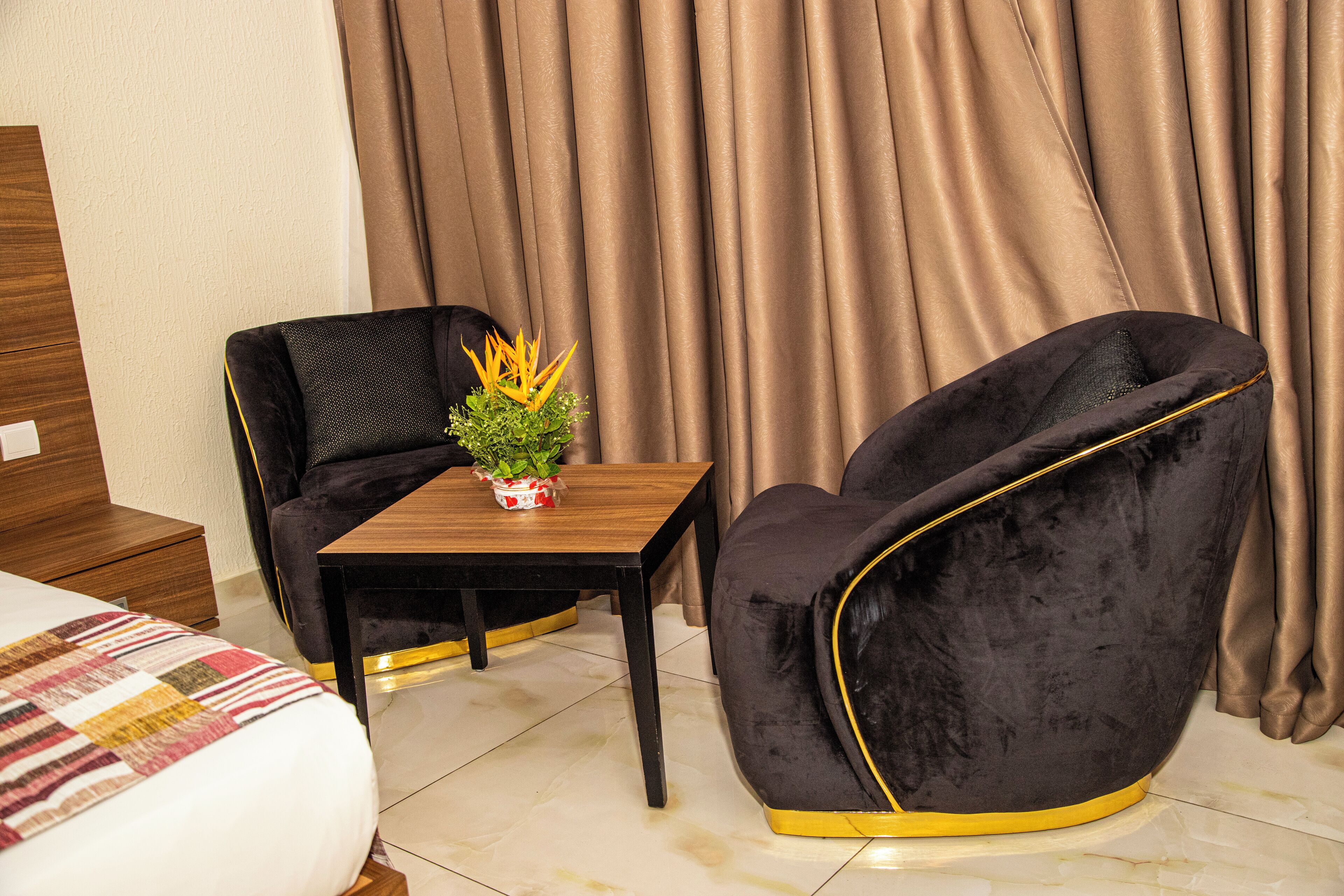 A hotel room with a bed, a small table with a plant, two black chairs, and a wooden nightstand.