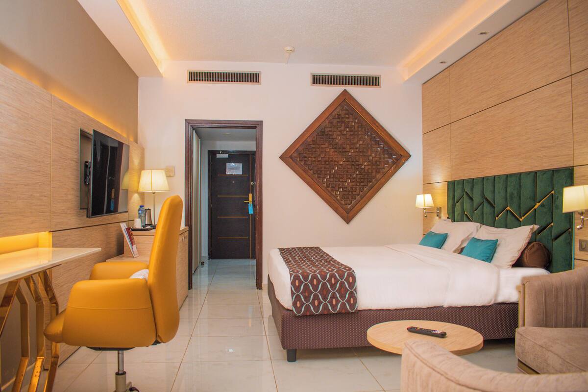 A hotel room with a bed, a desk, a chair, a small table, and a wall-mounted TV.