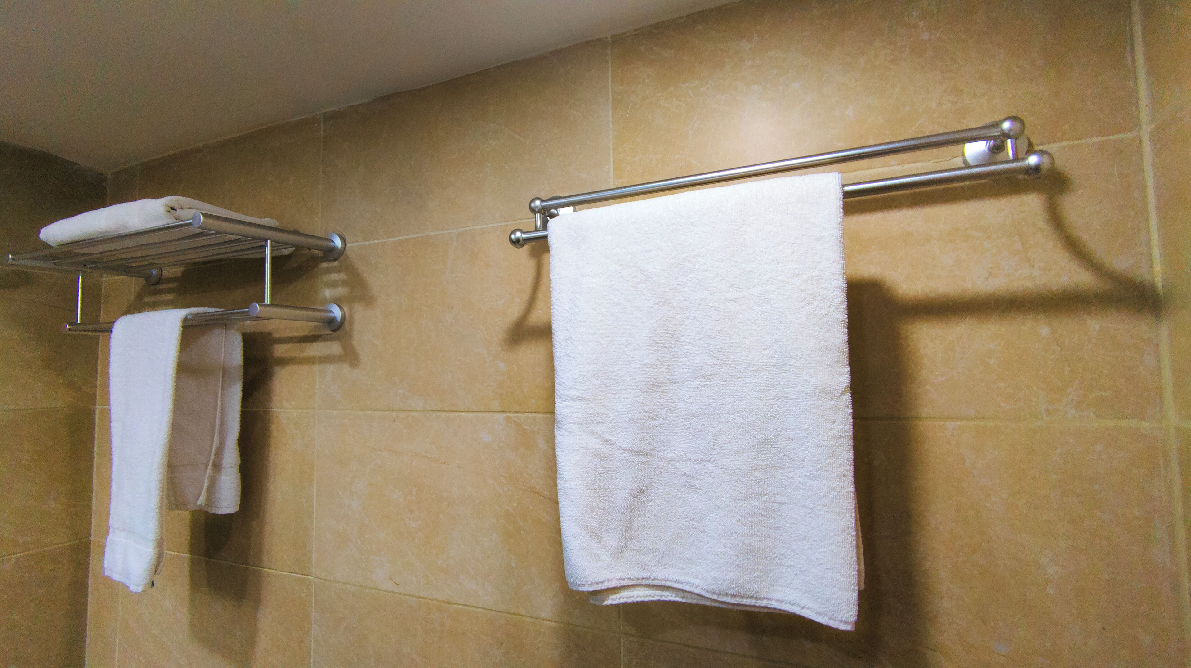 A bathroom with two white towels hanging on metal towel racks against beige tiled walls.