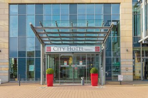 Entrada del Hotel City con fachada de vidrio, una cubierta metálica y macetas rojas.