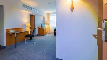 Habitación de hotel con escritorio, silla y lámpara. Se ve una puerta y una ventana con cortinas.