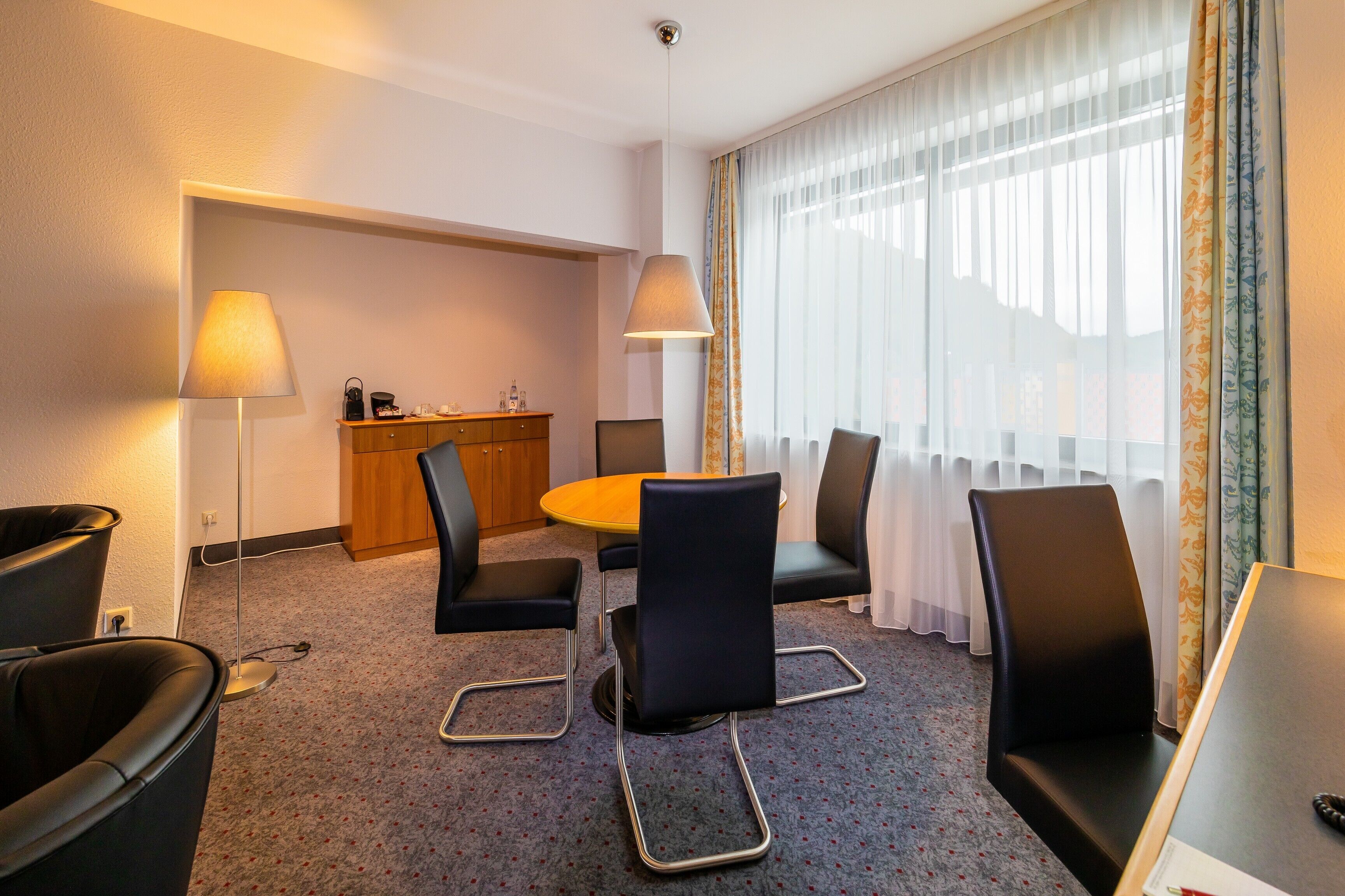 A hotel room with a round table, four chairs, a desk, and a lamp.