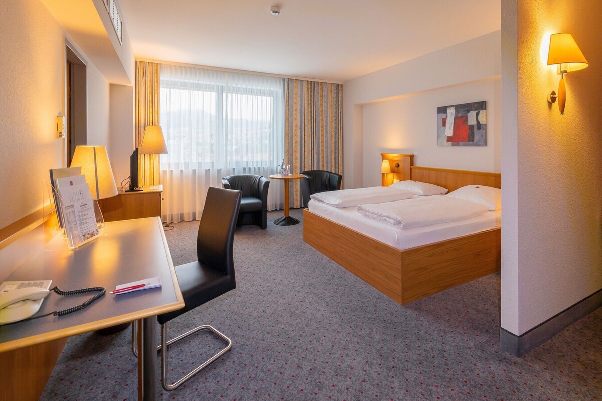 A hotel room with a bed, desk, chair, and a small table with a lamp.