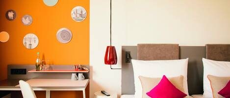 Una habitación de hotel con escritorio, cama con almohadas, lámpara roja y adornos en la pared.