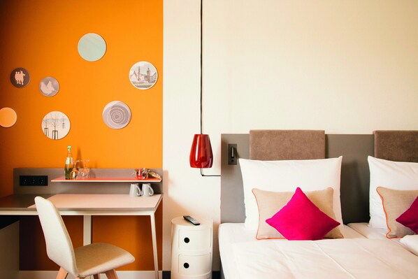 A hotel room with a desk, a bed with pillows, a red lamp, and wall decorations. - Vienna House Easy by Wyndham Wuppertal (Wuppertal)