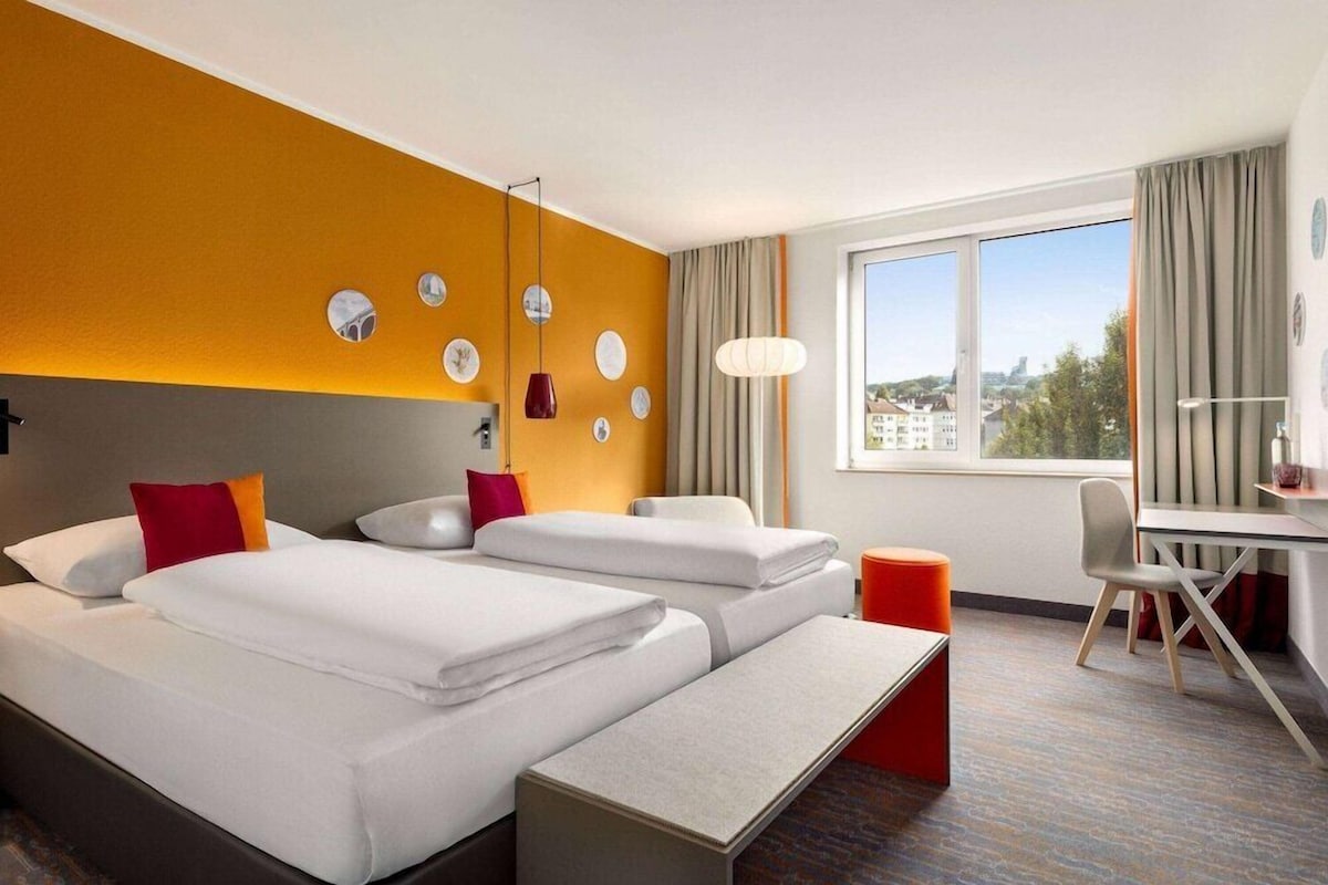 A hotel room with two beds, a desk, and a large window offering a city view.