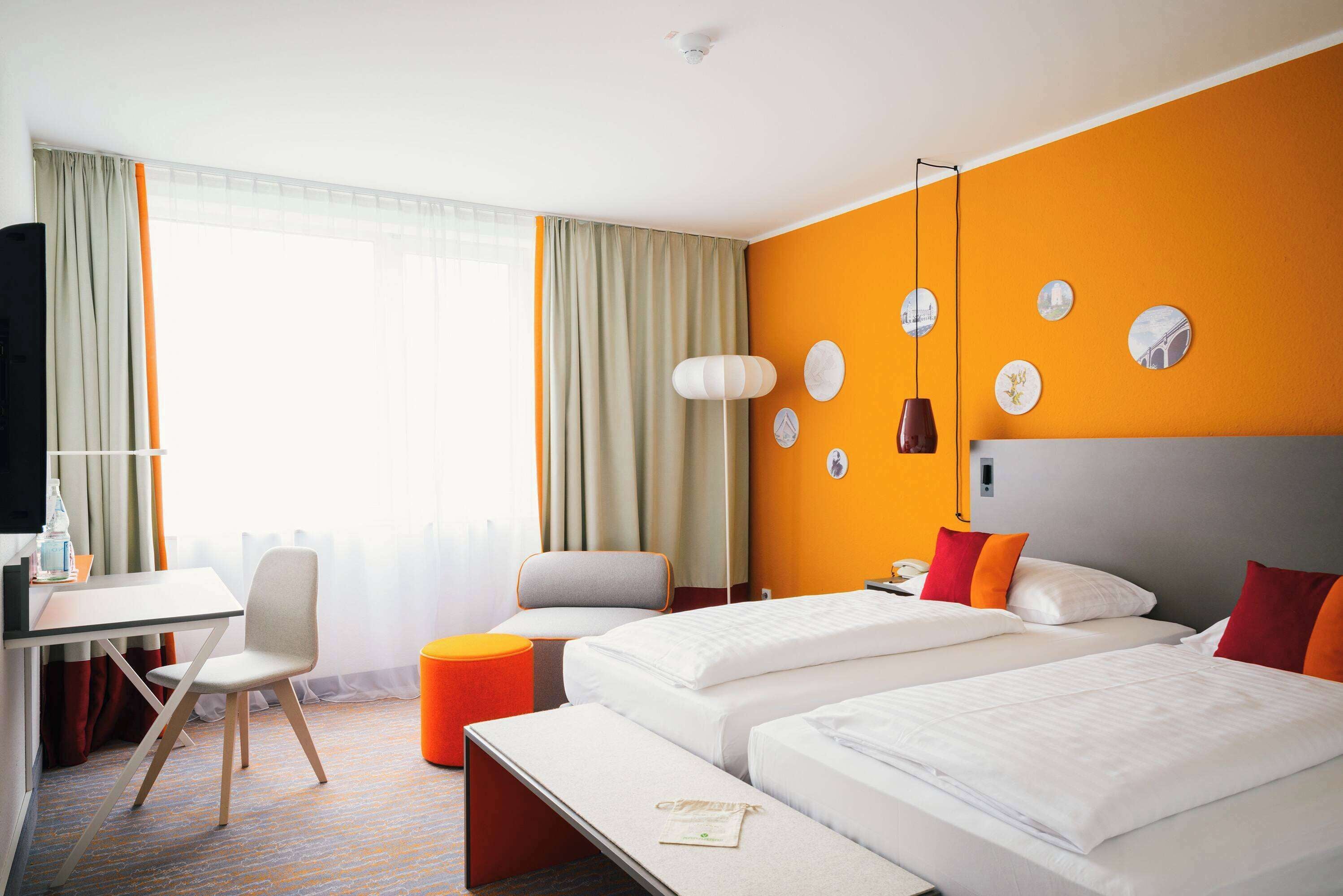 A hotel room with two beds, a desk, a chair, a small orange ottoman, and a large window with curtains.