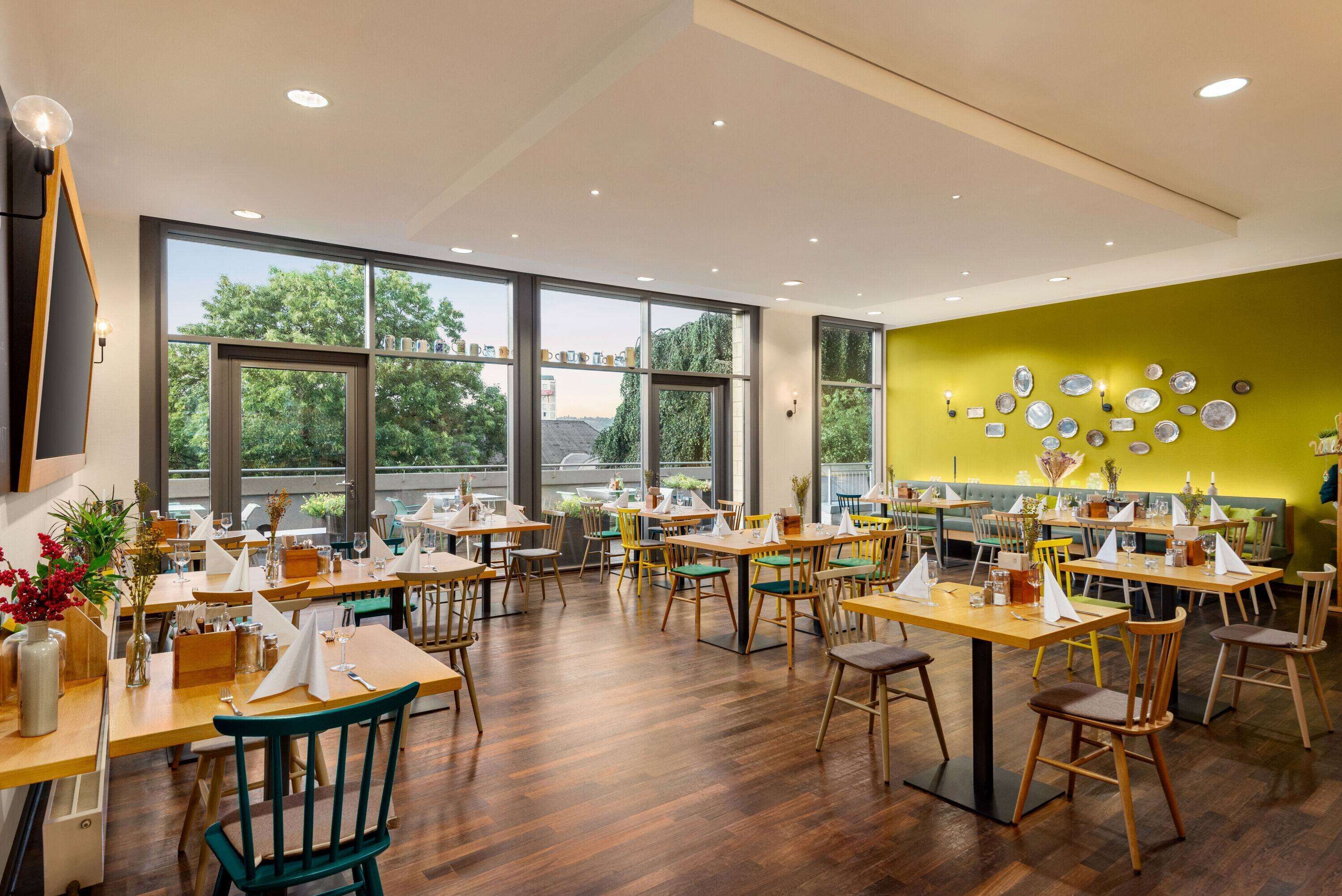 Un restaurante con mesas y sillas de madera, ventanas amplias y una pared verde decorada con platos.