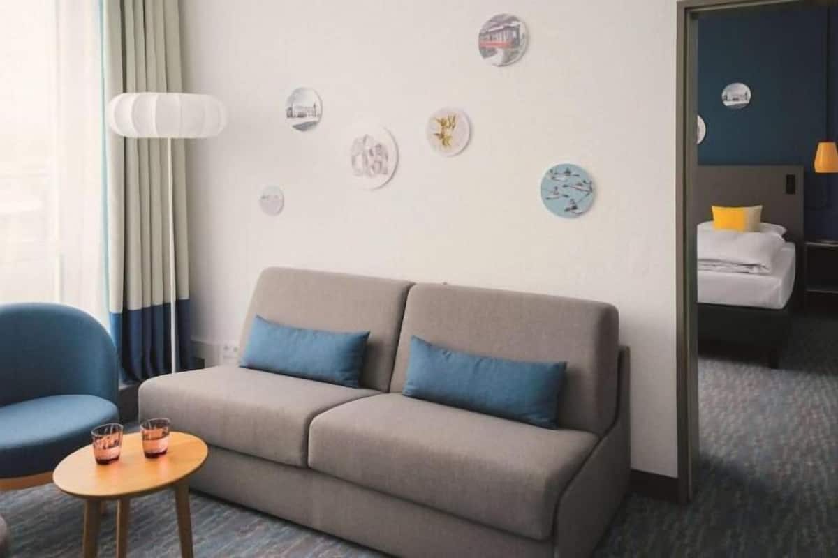 Habitación de hotel con sofá, una silla, una mesita con vasos y una cama con almohada.