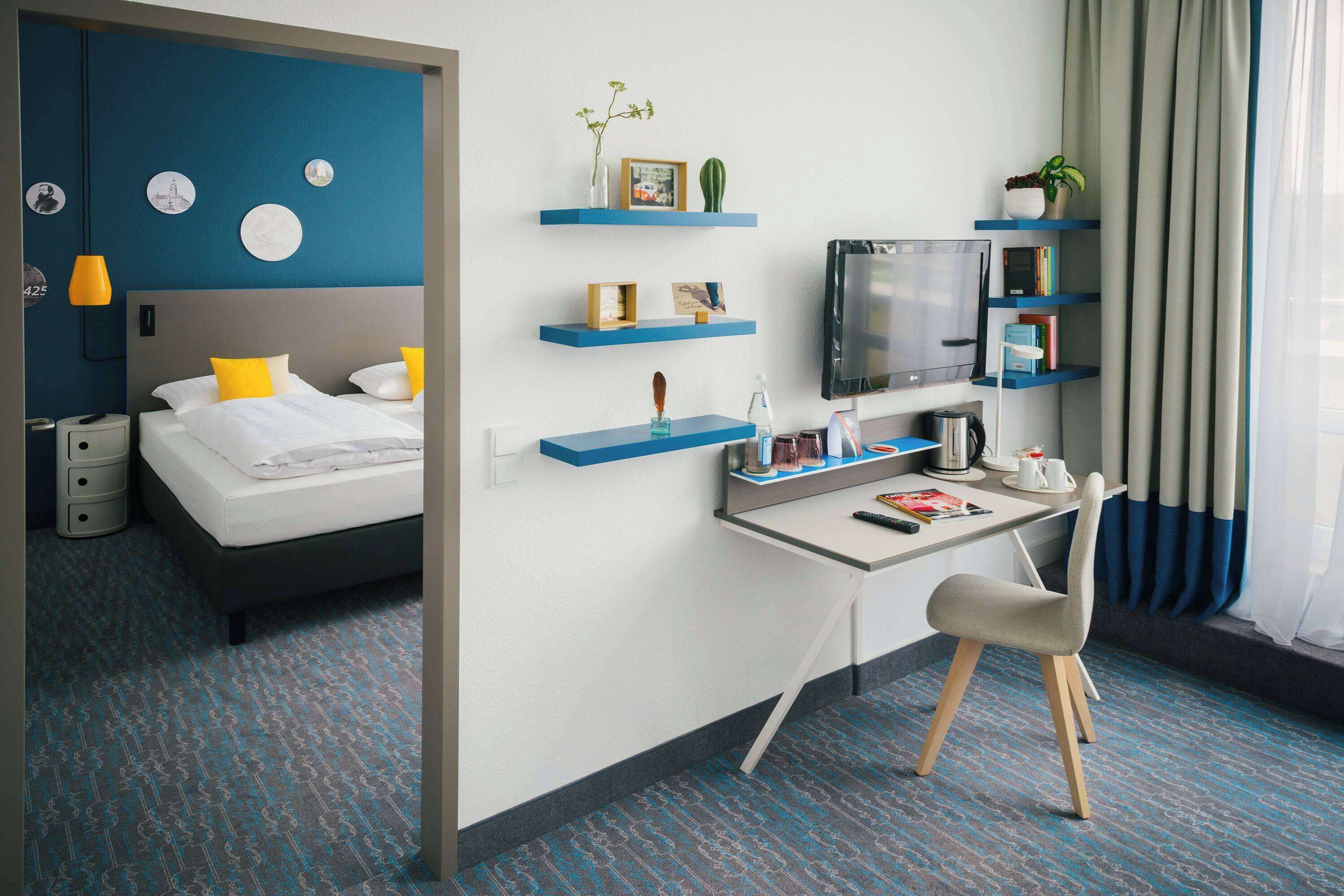 Ein modernes Hotelzimmer mit Bett, Schreibtisch mit Stuhl, Fernseher und Regalen mit Dekoration.