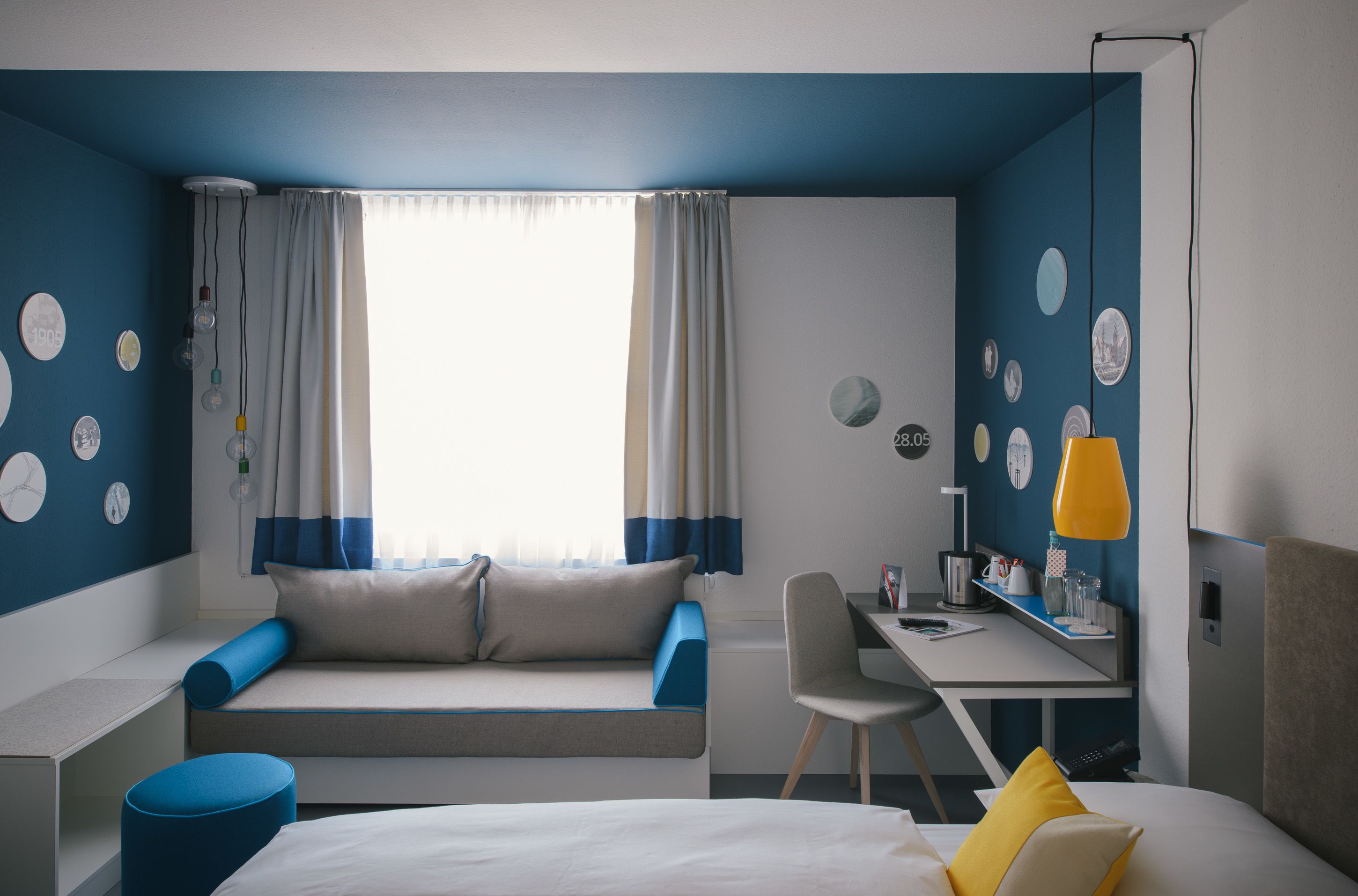 A modern hotel room with a bed, a desk, a chair, and a seating area with a sofa and curtains.