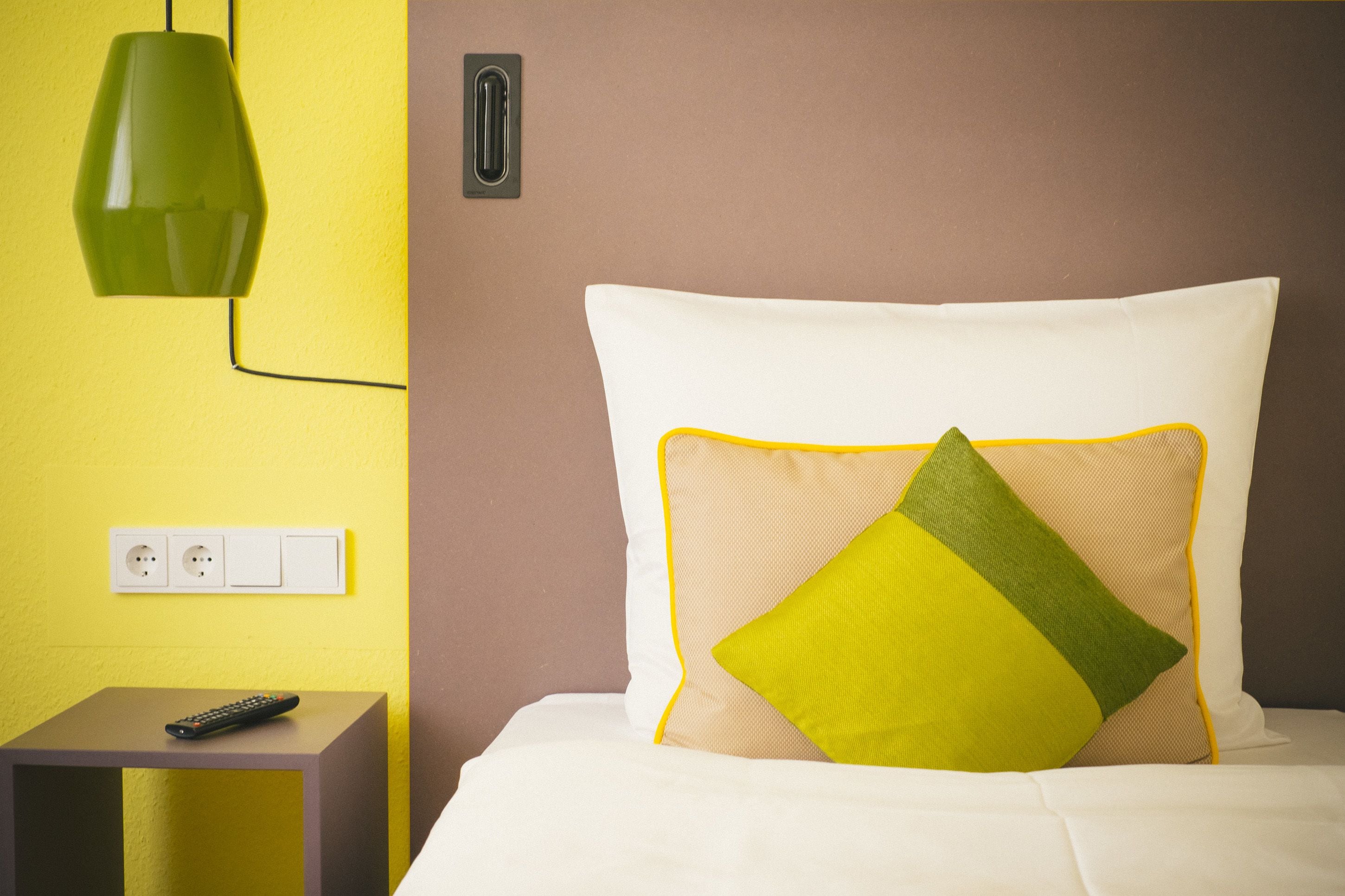 Una camera d'albergo con un letto, un comodino, una lampada a sospensione verde e una presa elettrica.