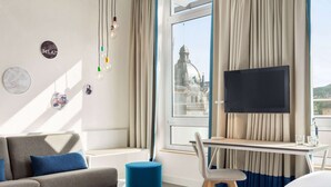 Una habitación de hotel moderna con sofá, reposapiés y un escritorio con silla. Un televisor de pantalla plana está montado en la pared, y hay cortinas en las ventanas.