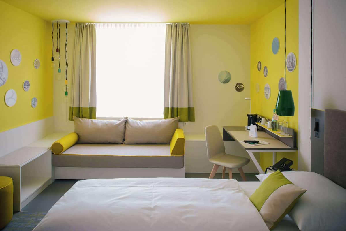 Una habitación de hotel con cama, escritorio, silla, sofá y ventana con cortinas.