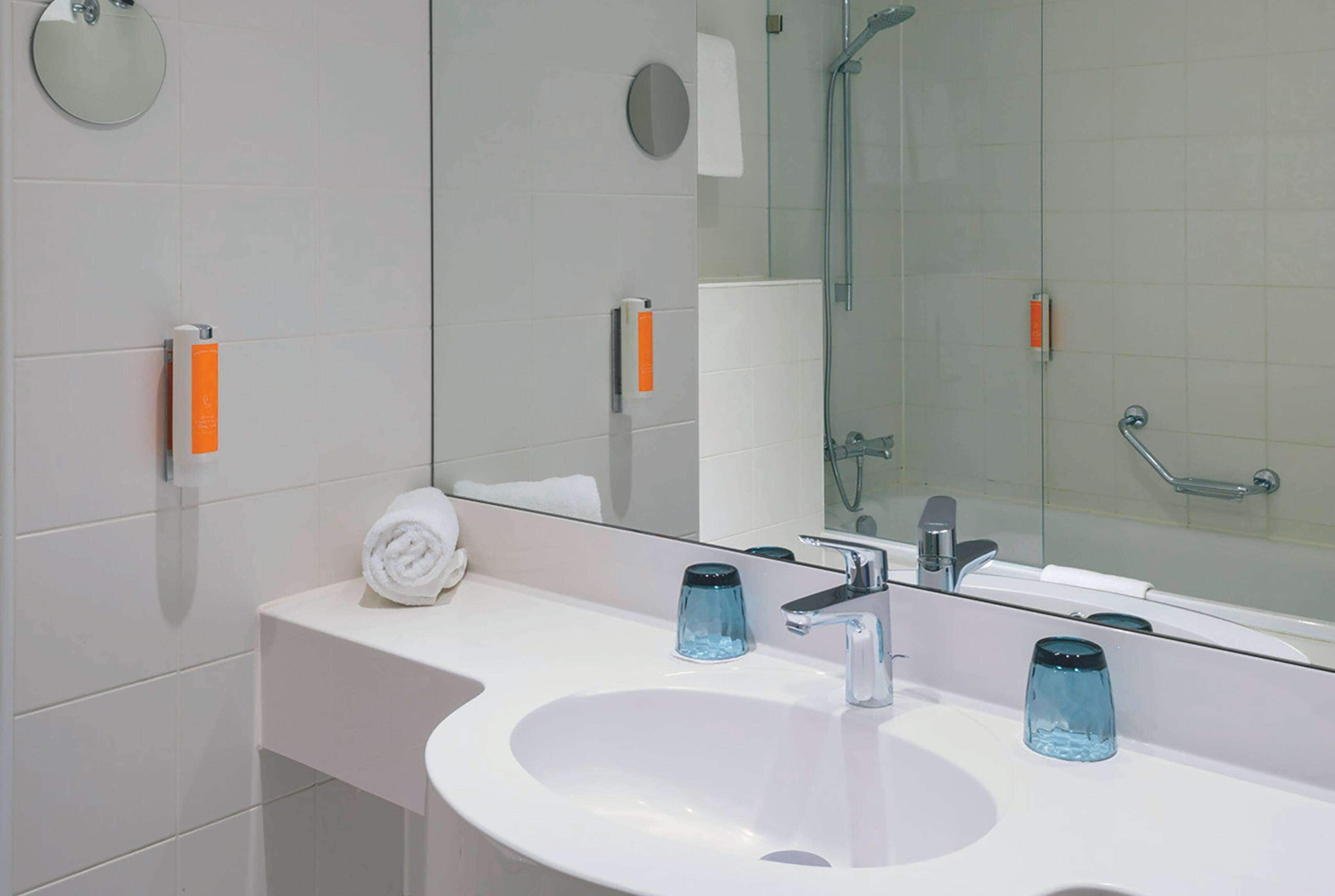 Ein modernes Badezimmer mit einem weißen Waschbecken, zwei blauen Glasbehältern, einem Handtuch und einem Spiegel, der einen Duschbereich reflektiert.