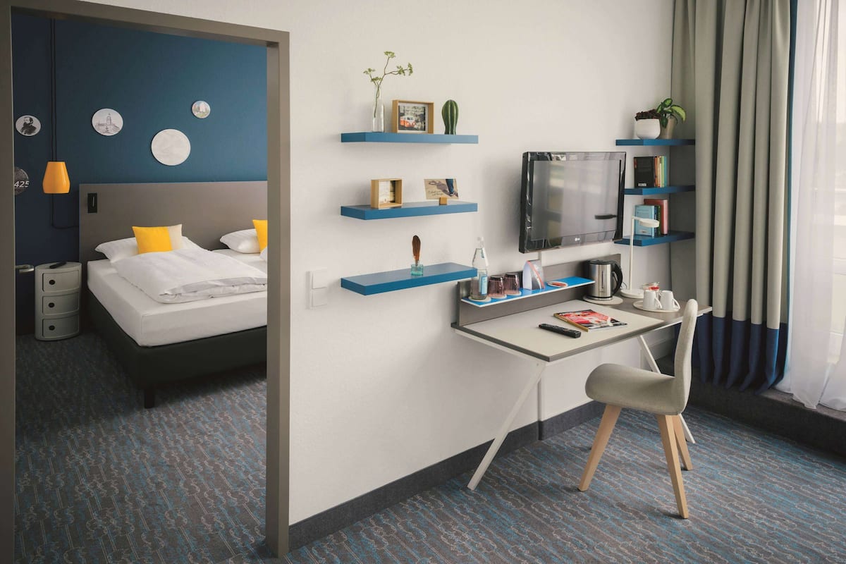 Una habitación de hotel moderna con una cama, un escritorio, una silla, un televisor y estantes con objetos decorativos.