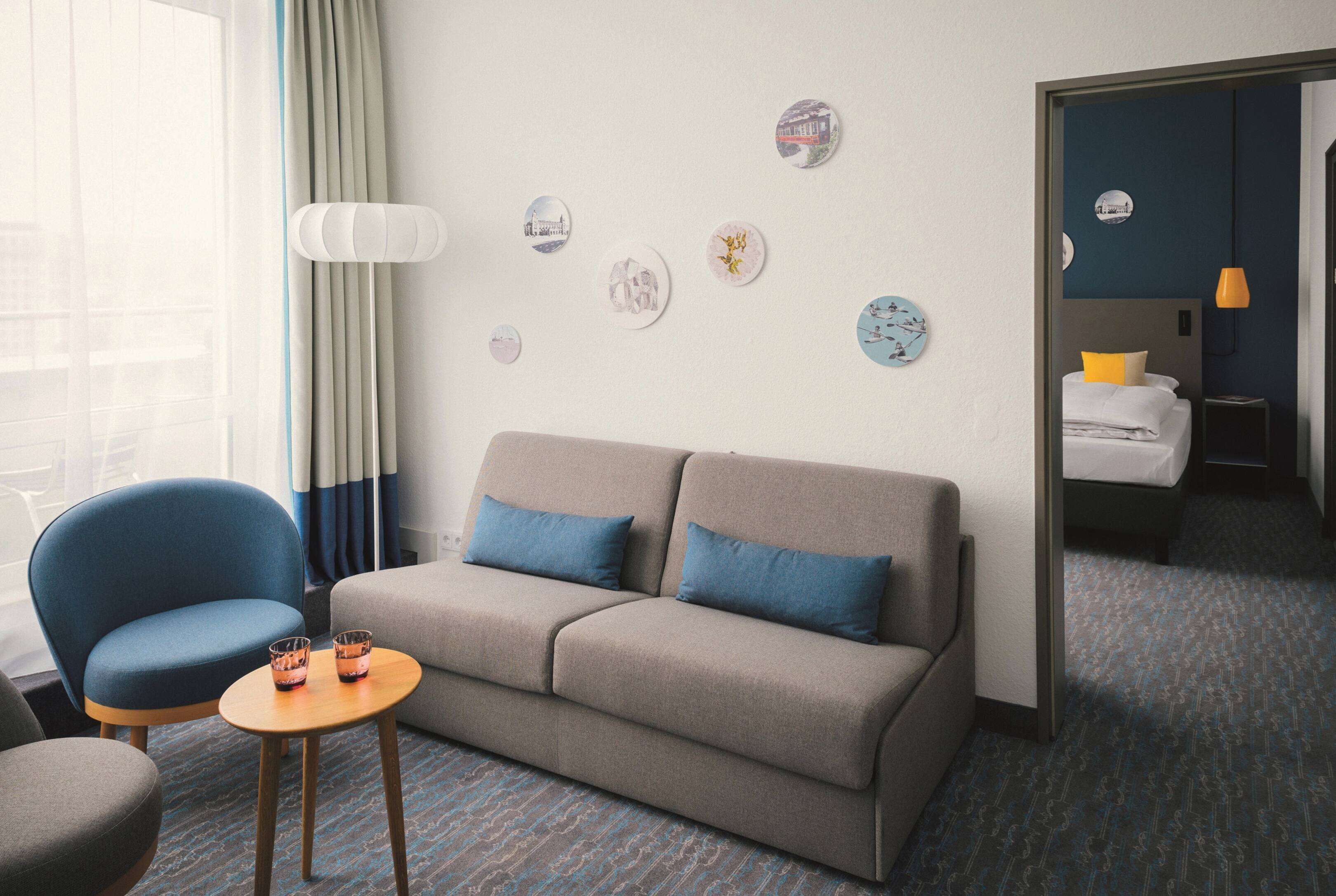 Habitación de hotel con sofá, dos sillones, una mesita con vasos y una cama con almohada amarilla.