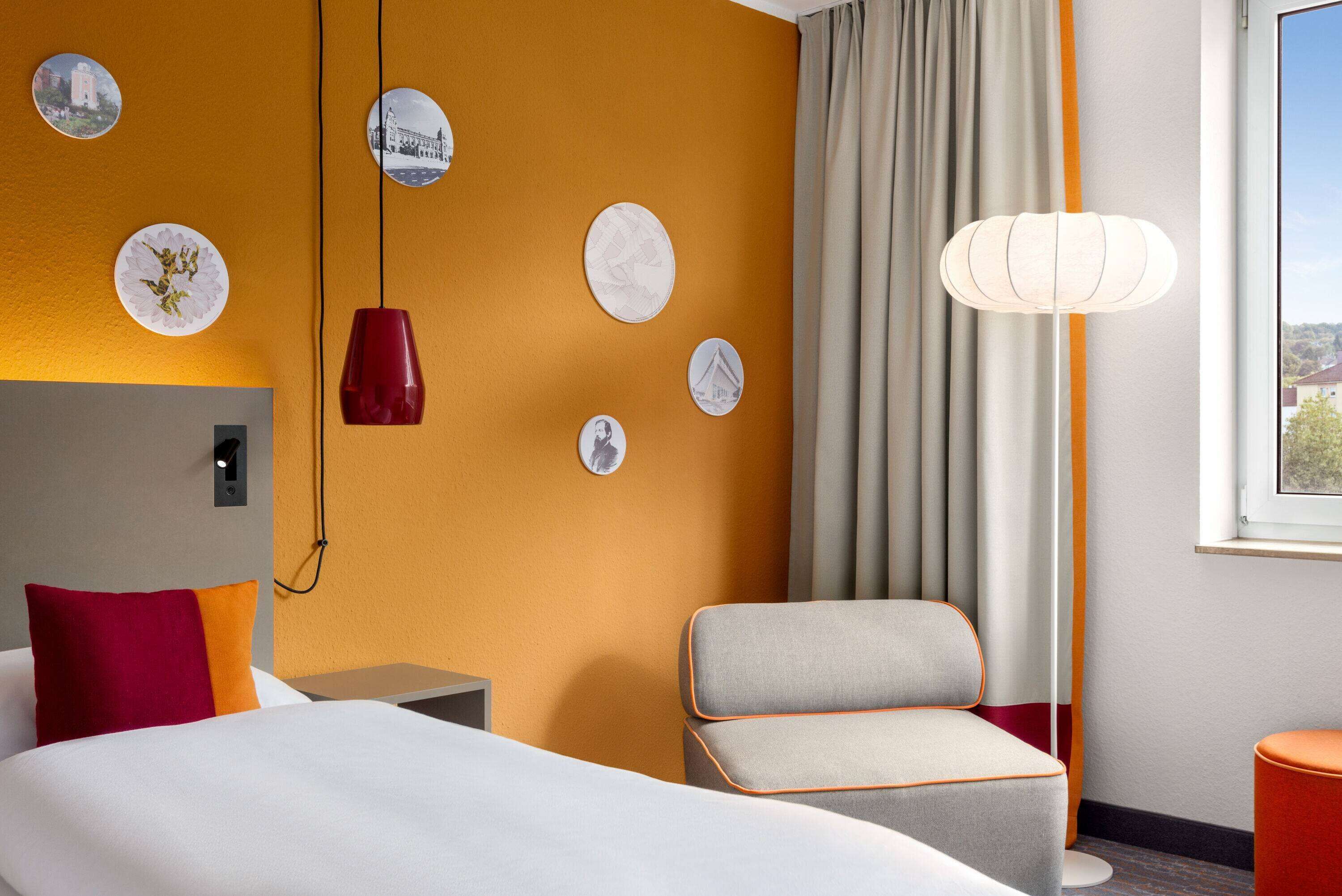 Una habitación de hotel con una cama, una silla, un escritorio, una lámpara y decoración en la pared.