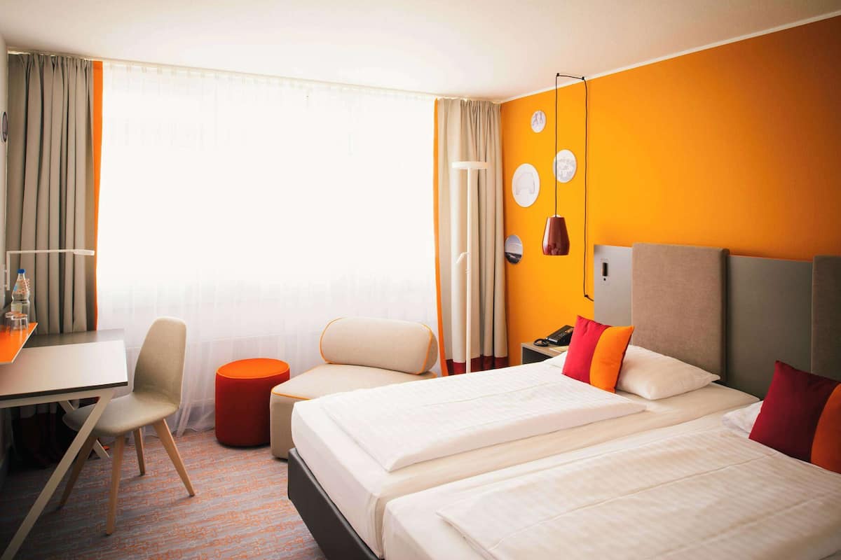 Habitación de hotel con dos camas, un escritorio, una silla y un pequeño taburete naranja.