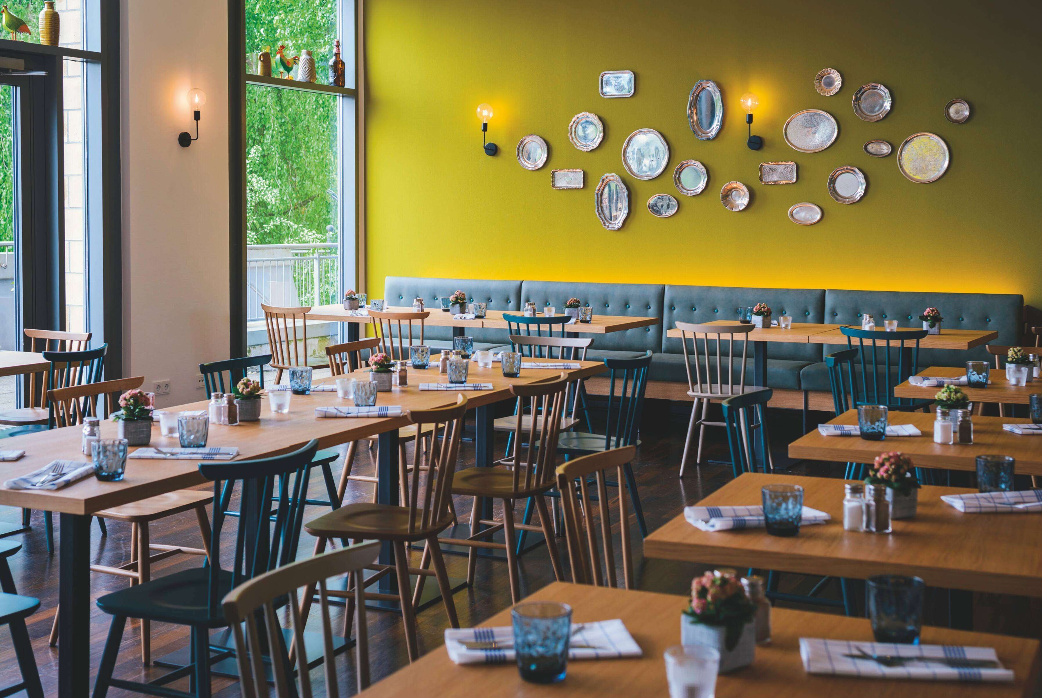 Un restaurant amb diverses taules i caires, una paret groga amb plats decoratius, i una banca verda amb coixins.