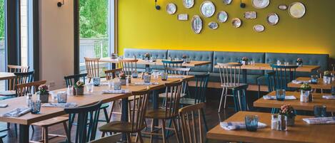 Un restaurante con varias mesas y sillas, una pared amarilla adornada con platos decorativos y un banco acolchado verde.