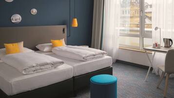 Habitación de hotel con dos camas, un escritorio, una silla, un taburete azul y una ventana con vistas a edificios.