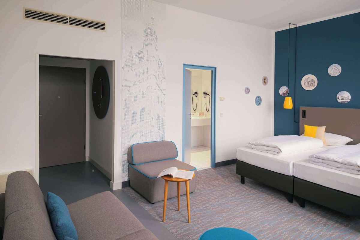 Habitación de hotel moderna con un sofá gris, un taburete de madera y una cama con ropa blanca, además de una pared azul de acento.