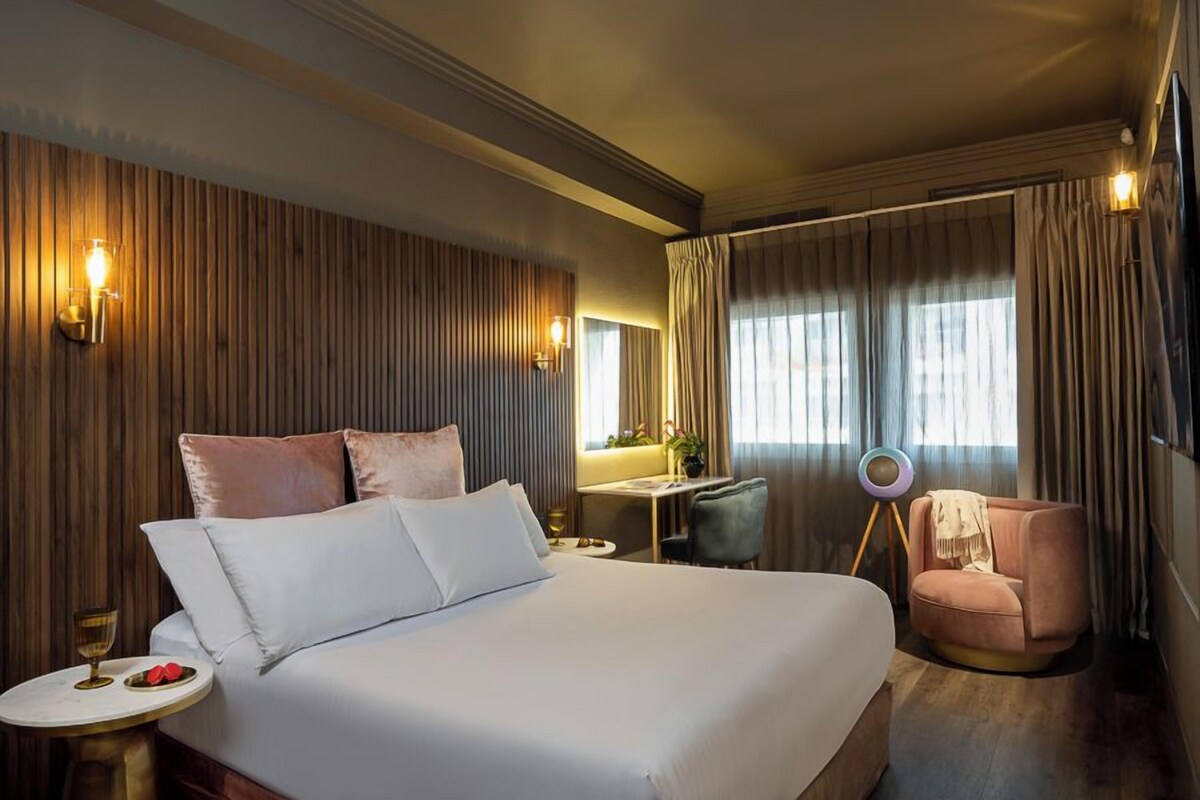 A hotel room with a large bed, a desk, a chair, and a small table with a glass and two red items.