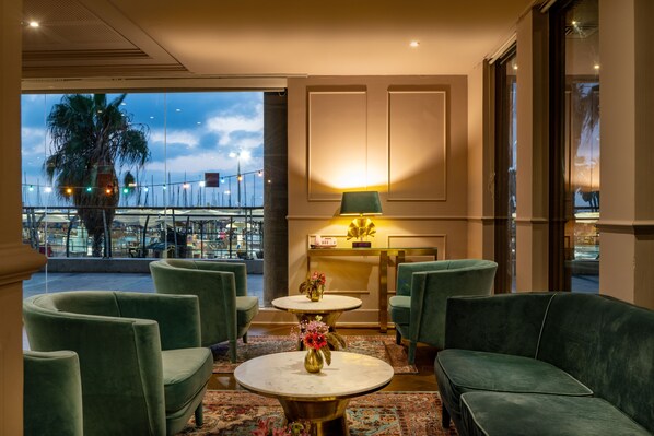 A lobby with green upholstered seating, a marble table with a vase of flowers, and a view of palm trees and a cityscape through large windows. - Leonardo Gordon Beach (Tel Aviv)