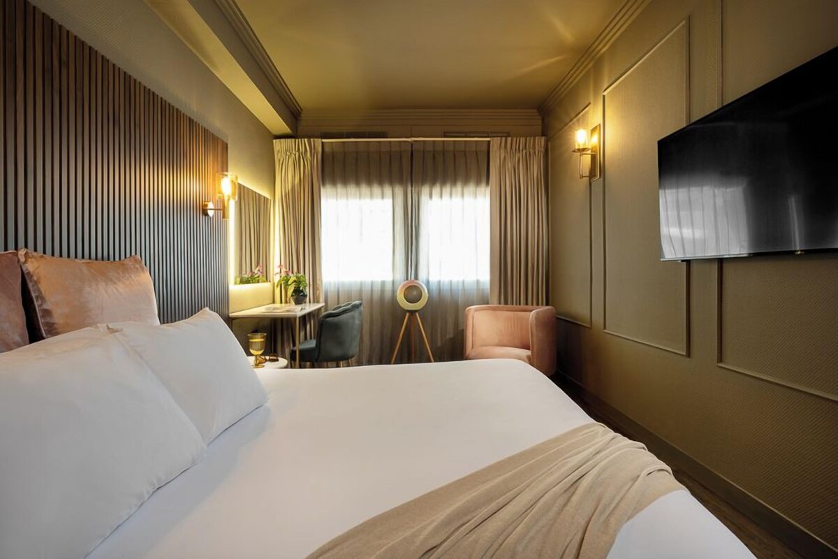 Habitación de hotel con una cama grande, un escritorio con silla, una mesa redonda con lámpara y un televisor montado en la pared.