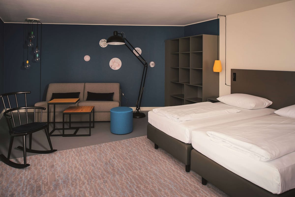 A hotel room with a bed, a sofa, a desk, a chair, and a blue ottoman.