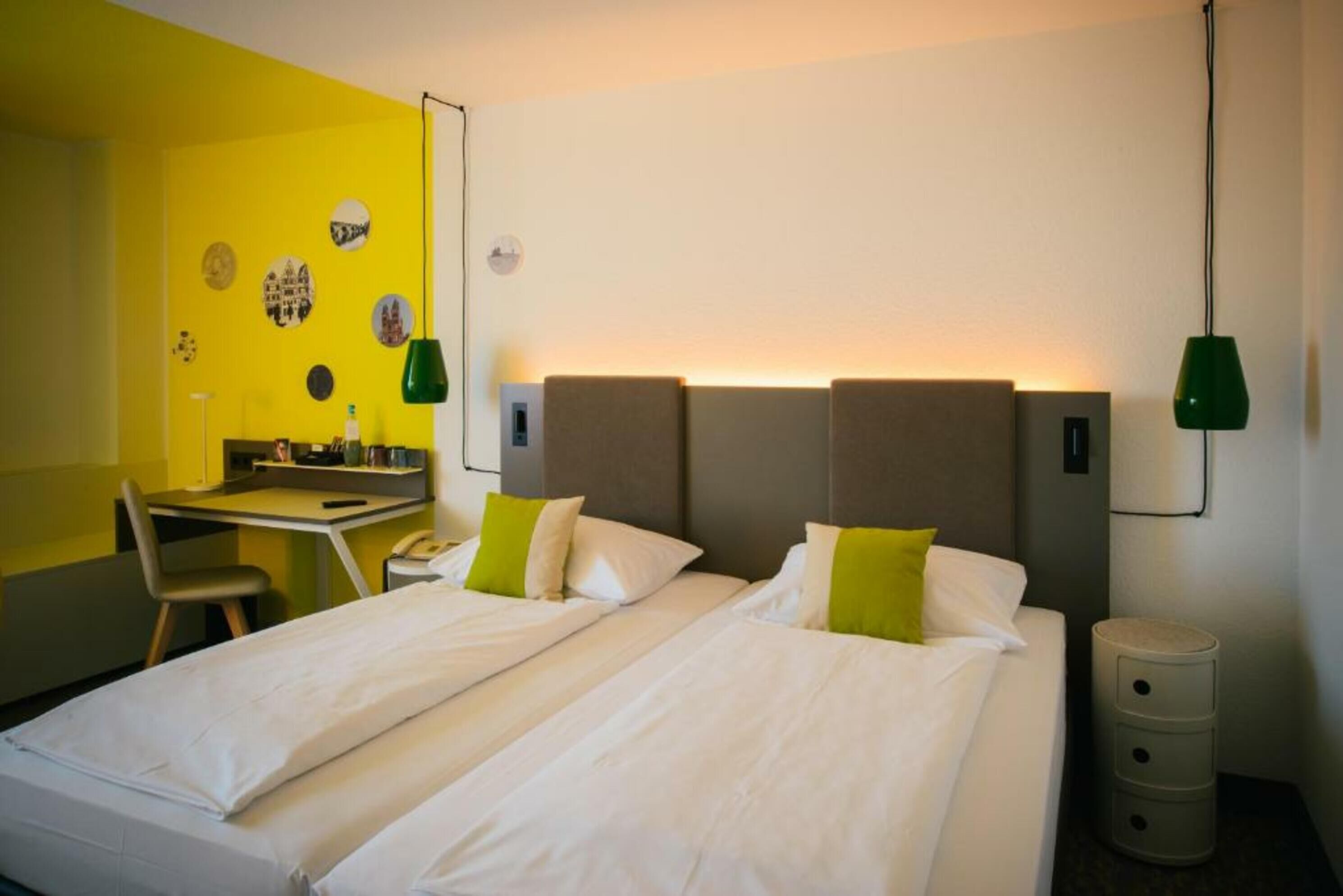 Habitación de hotel con dos camas, un escritorio y una silla. Hay lámparas fijadas a la pared y una mesita de noche con cajones.
