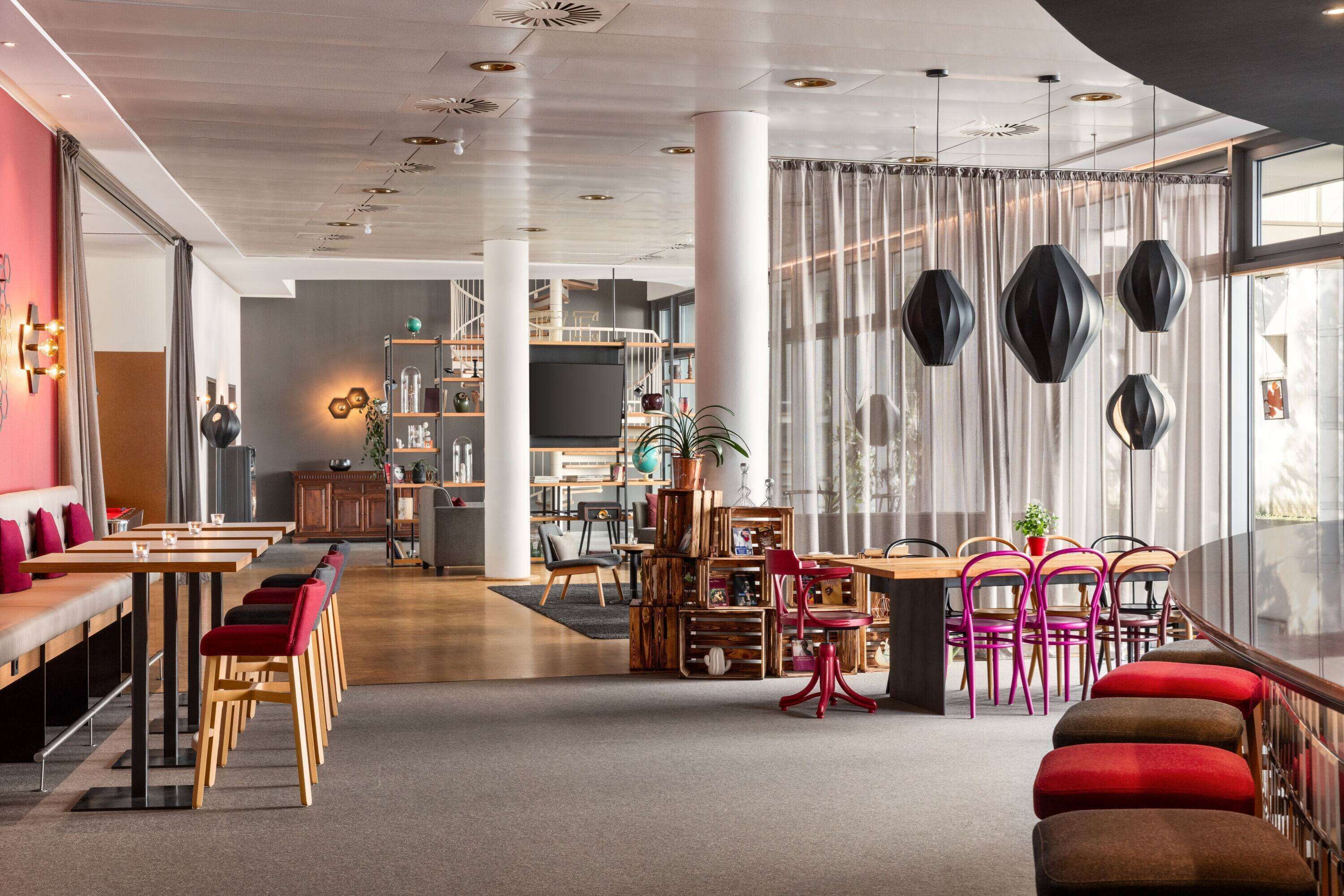 A modern interior with a long dining table, red and brown upholstered chairs, and a large window with sheer curtains.