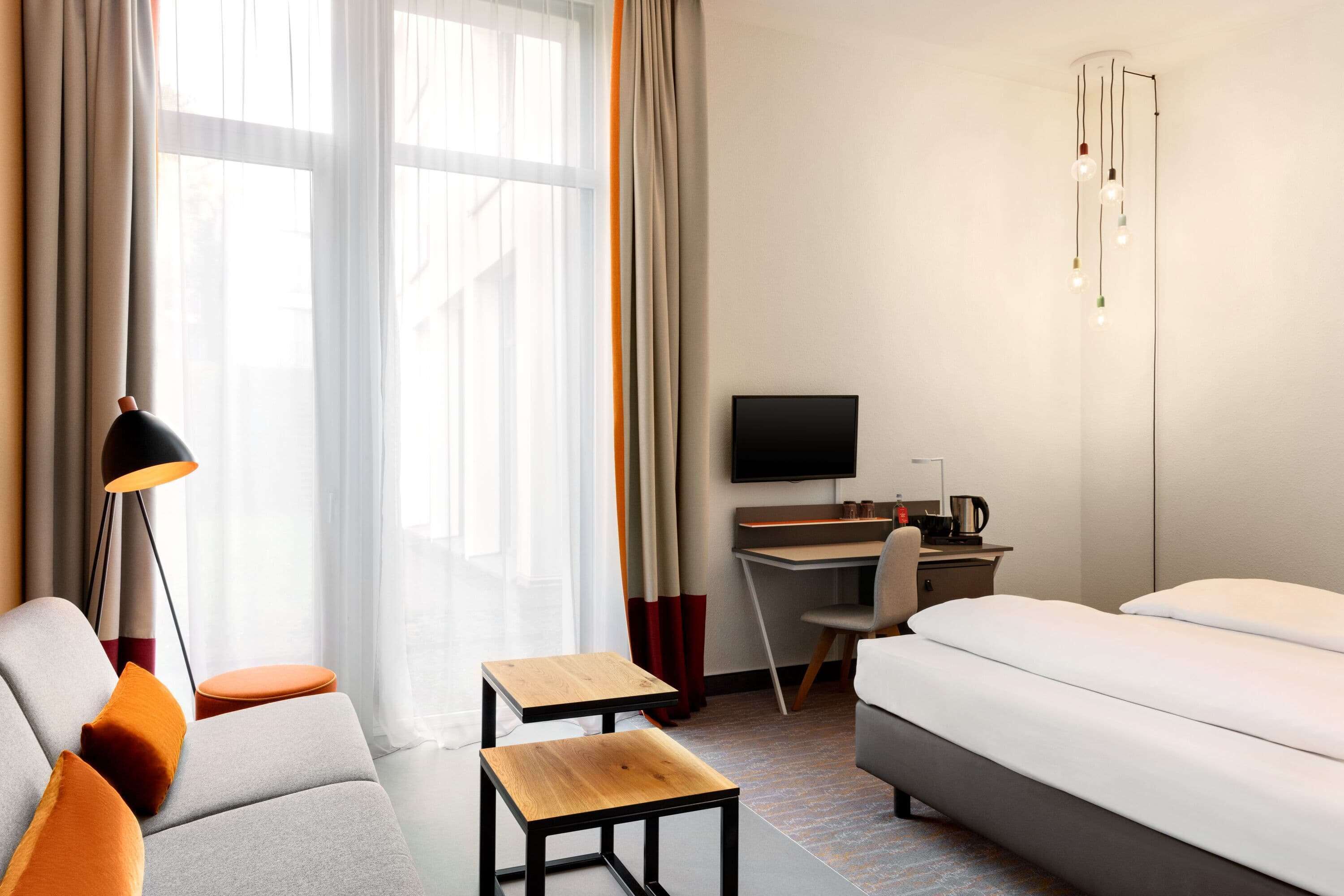Una habitación de hotel moderna con sofá, una mesita de madera, cama, escritorio con silla, televisor y una lámpara de diseño original.
