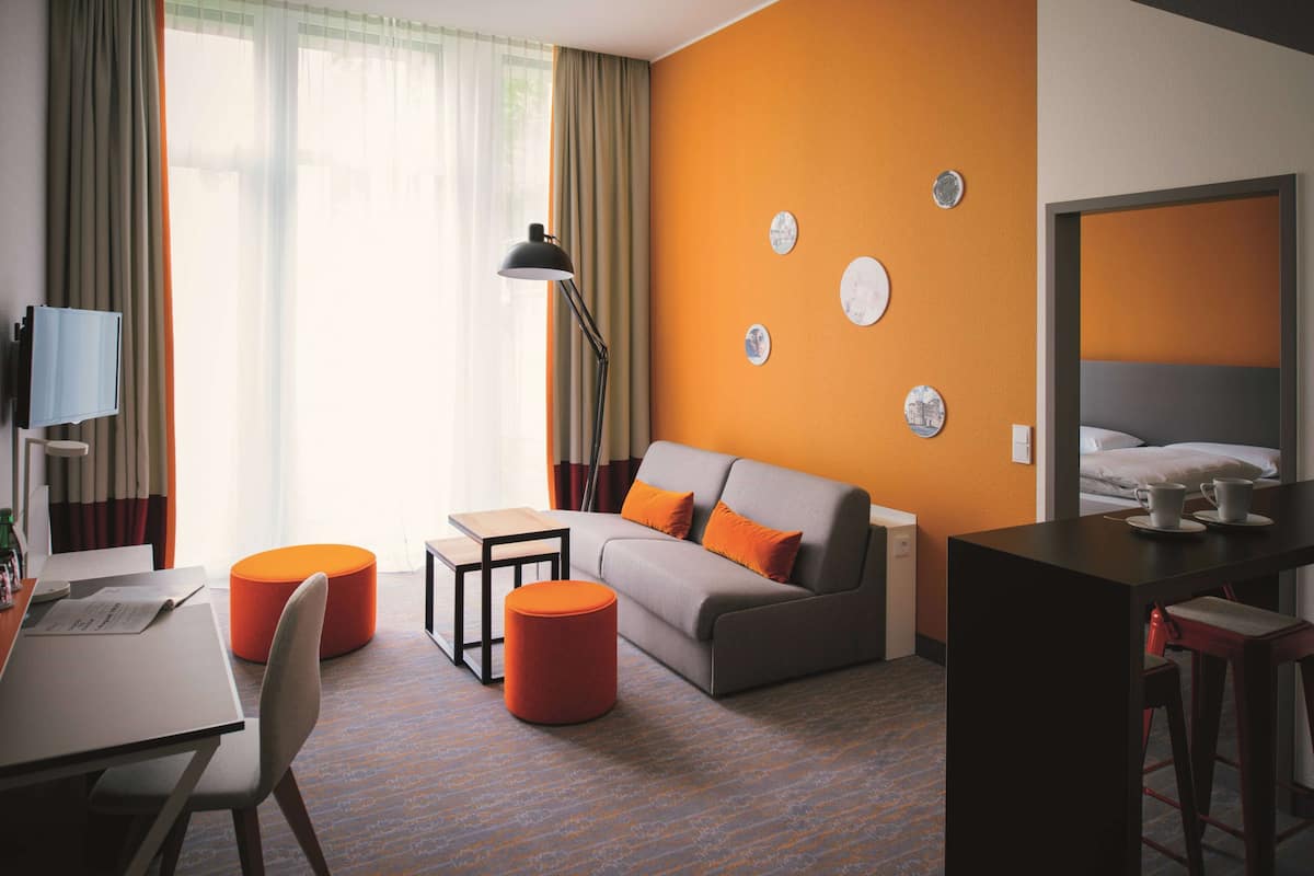 A modern hotel room with a sofa, ottoman, and a desk with a lamp.