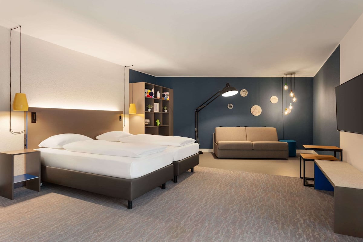 A modern hotel room with a bed, a sofa, a desk, and a bookshelf.