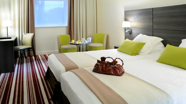 Une chambre d’hôtel avec deux lits, un bureau, une chaise, une petite table avec des articles pour le petit-déjeuner et une fenêtre avec des rideaux.