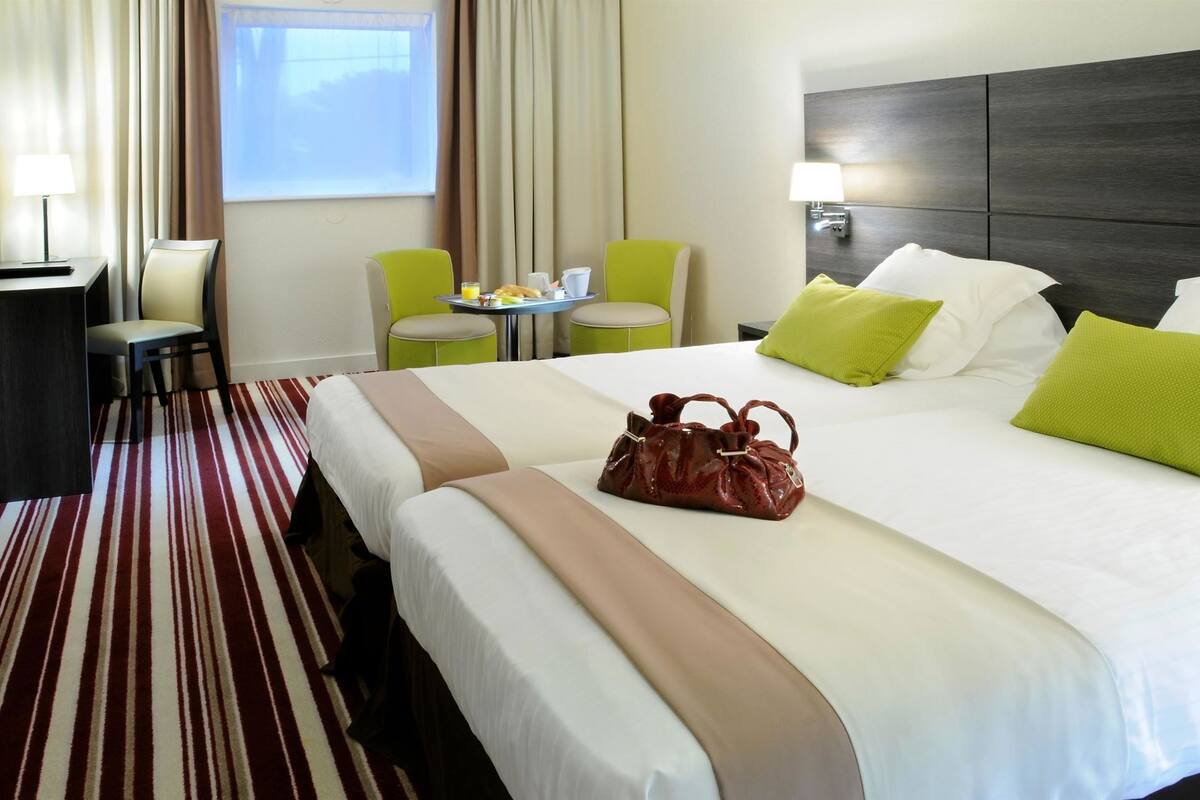 A hotel room with a bed, a desk, two chairs, a small table with breakfast items, and a window with curtains.