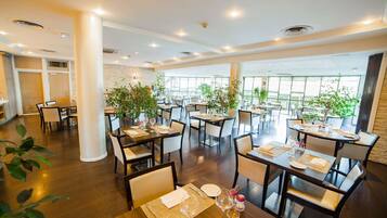 Un vaste espace dédié à la salle à manger, avec de nombreuses tables et chaises, de grandes fenêtres et des plantes d’intérieur.