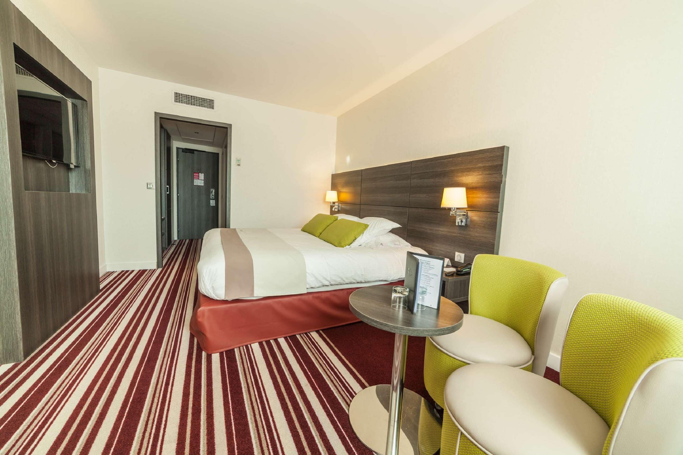 A hotel room with a bed, a small table, a chair, and a TV mounted on the wall.