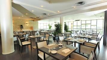 En rummelig restaurant med flere borde og stole, store vinduer og en stenvæg.