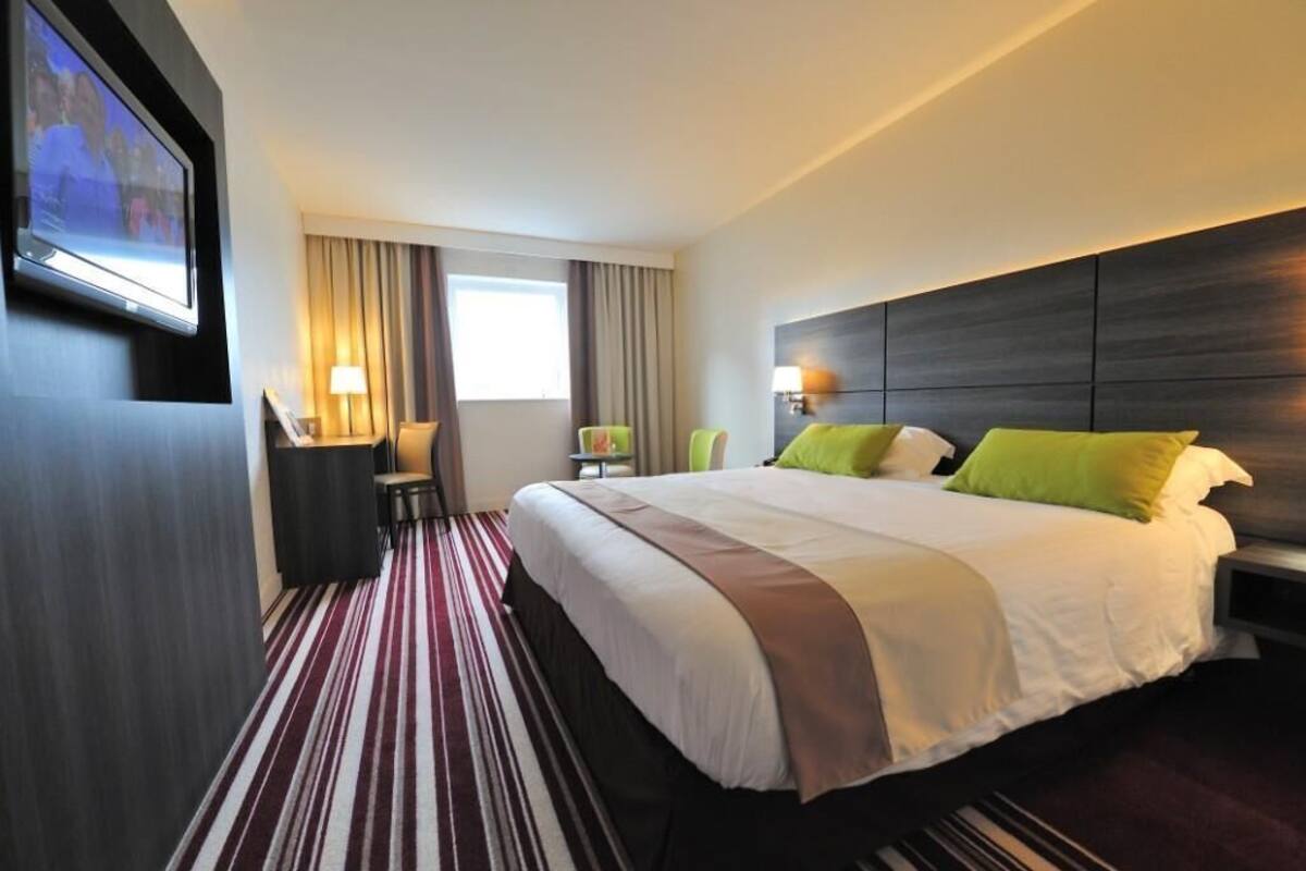 A hotel room with a large bed, a TV mounted on the wall, a desk with a chair, and a window with curtains.
