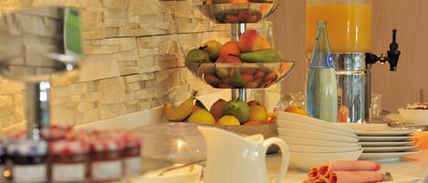 A buffet with a variety of fruits and pastries, a juice dispenser, and a stone wall in the background.