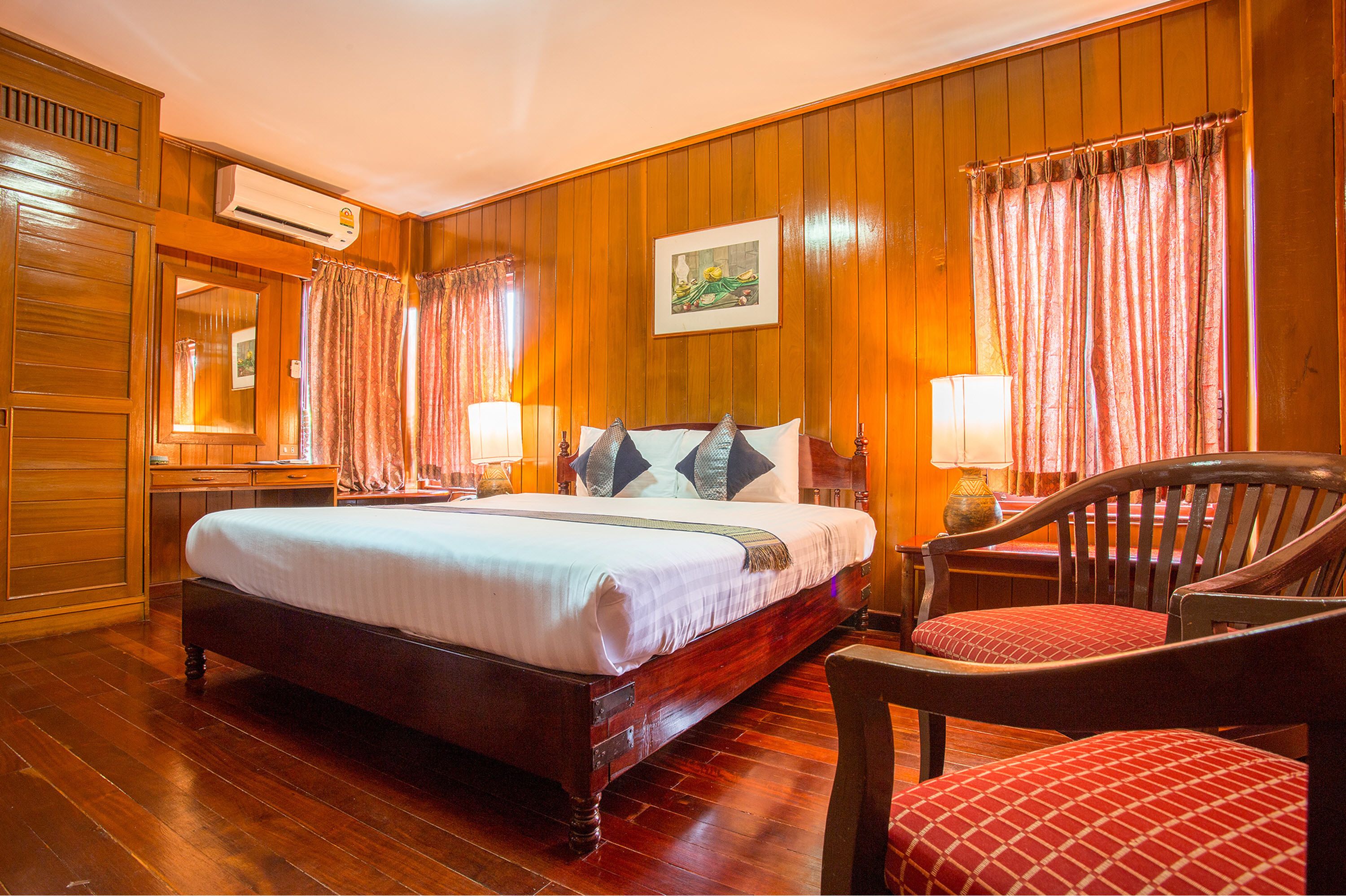Ein Hotelzimmer mit einer Wandpaneele aus Holz, einem großen Bett mit weißer Bettdecke, zwei Nachttischlampen, einem Holzsessel mit rotem Kissen und einem gerahmten Bild an der Wand.