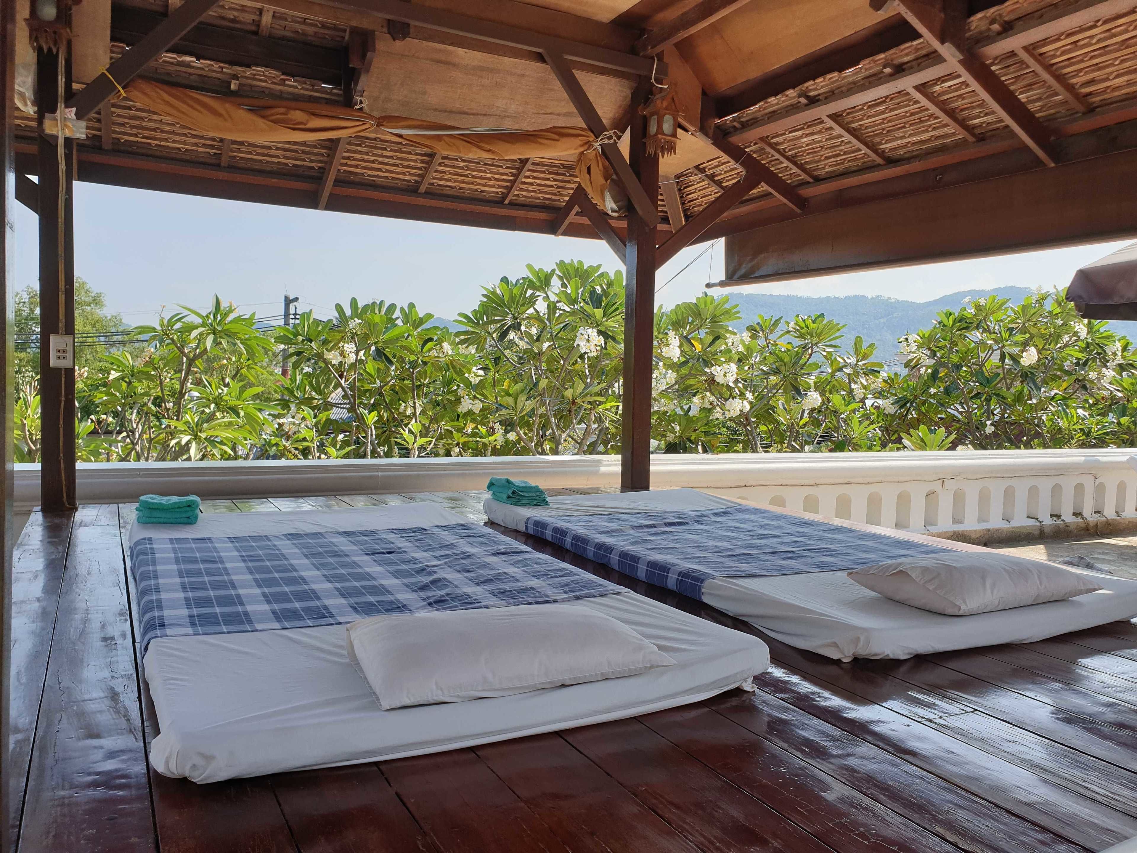 Terraza de madera con dos camas, cada una con colcha azul a cuadros y almohadas blancas, con vistas a un jardín exuberante y montañas lejanas.